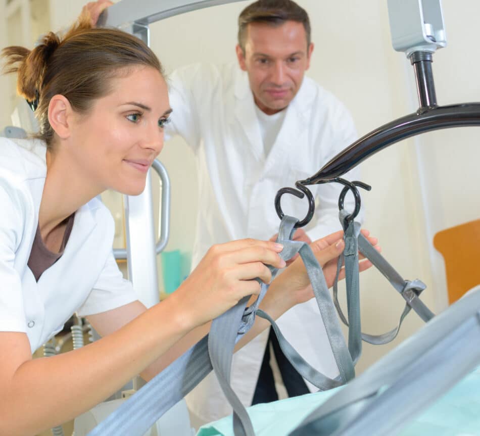 Two healthcare workers in uniforms adjust a medical hoist near a hospital bed.