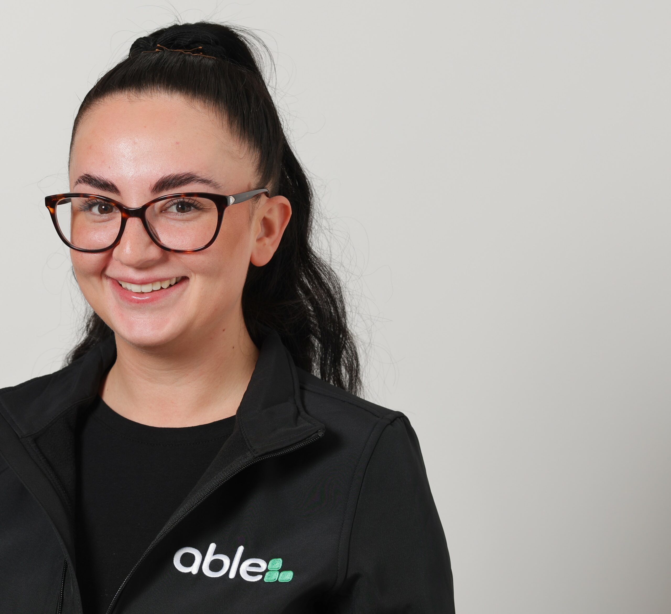 Person with glasses and a ponytail, wearing a black jacket with an "able" logo, smiles at the camera against a plain background.