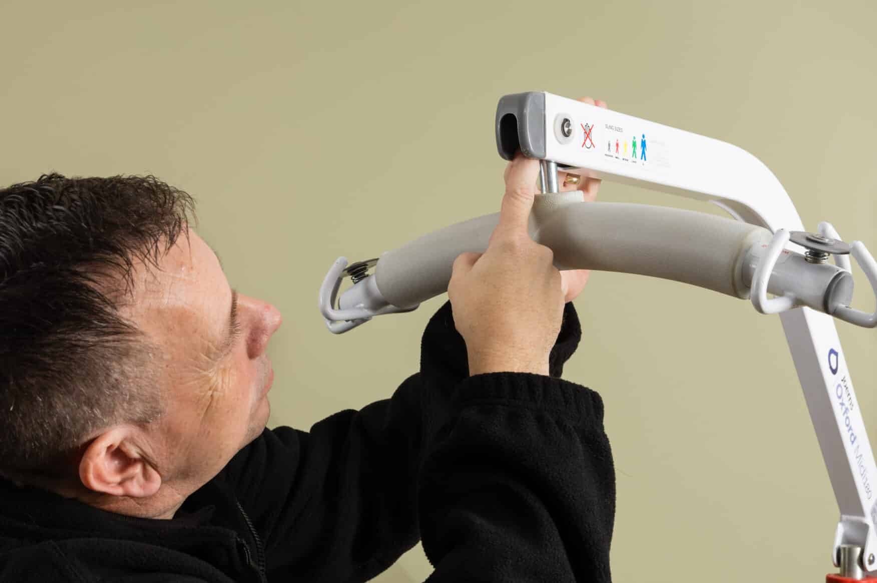 A man adjusts or completes a care home loler and adjusts the control panel of a patient lifting device against a plain beige background.