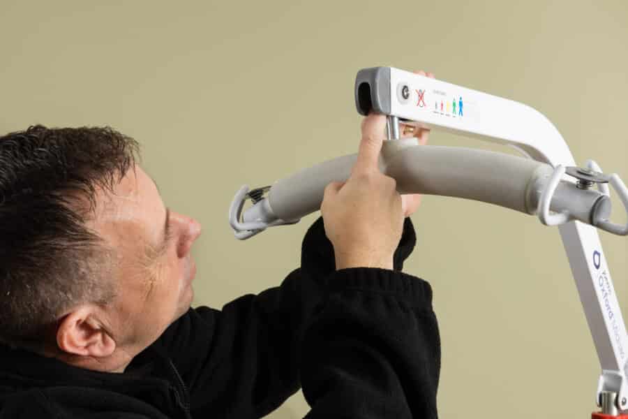 A man adjusts or completes a care home loler and adjusts the control panel of a patient lifting device against a plain beige background.