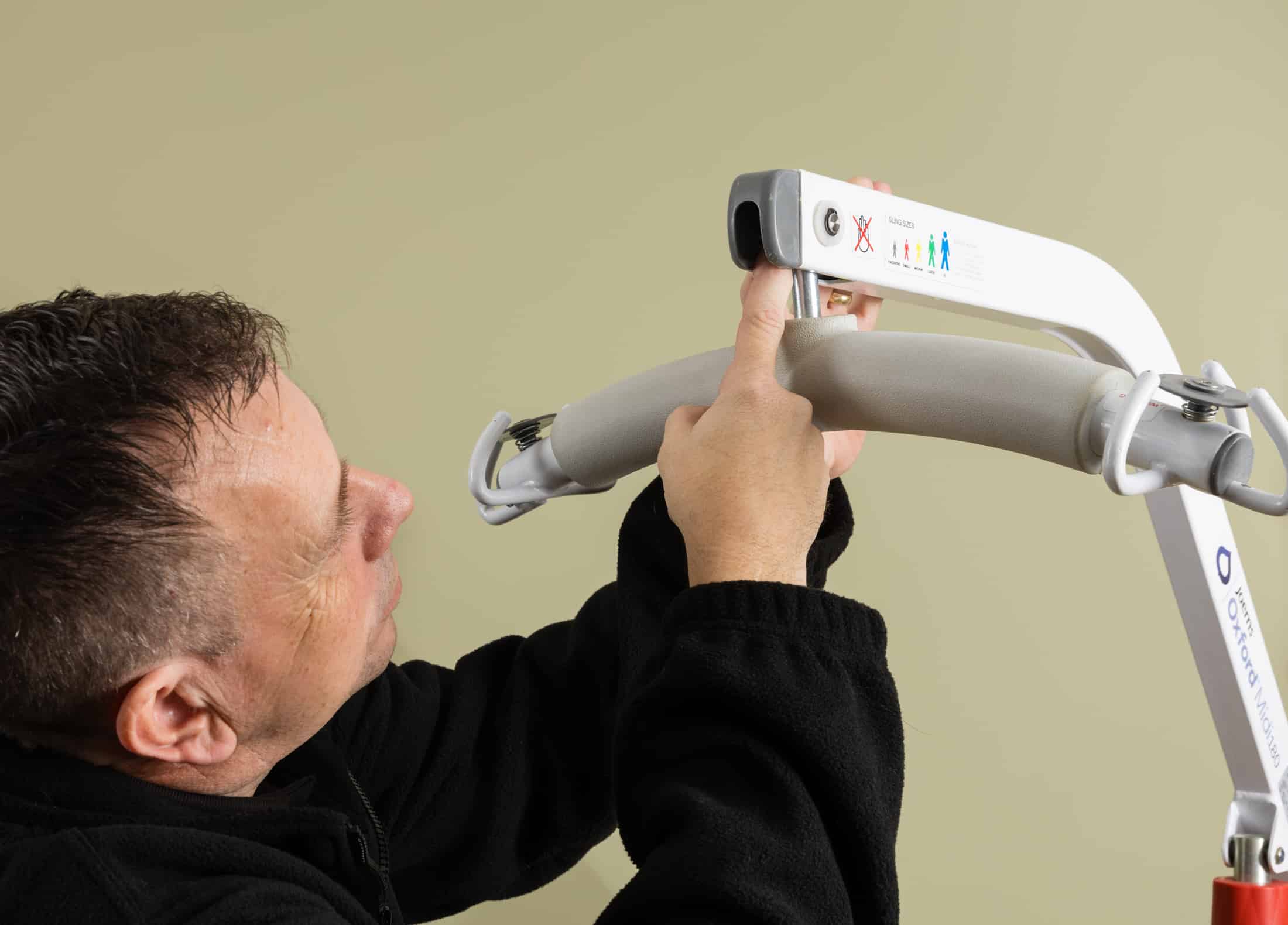 A man adjusts or completes a care home loler and adjusts the control panel of a patient lifting device against a plain beige background.