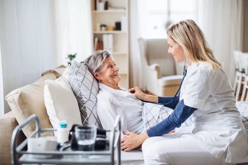 A nurse in uniform checks on an elderly woman lying in fire retardant bedding in a care home, both smiling, with medical supplies on a nearby table.