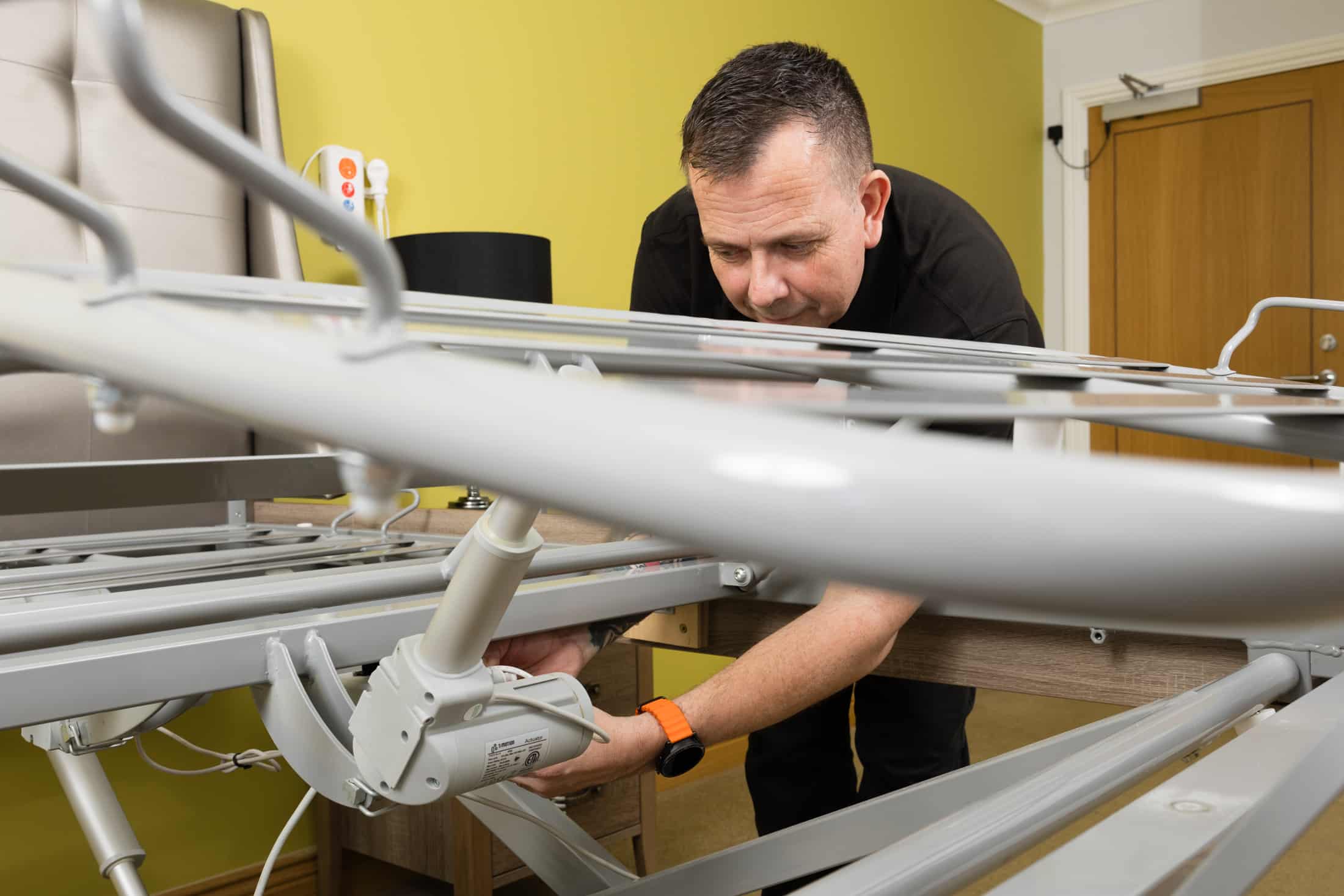 A man, who can carry out a loler test, adjusts the mechanism of a metal hospital bed frame in a room with yellow walls and a wooden door.