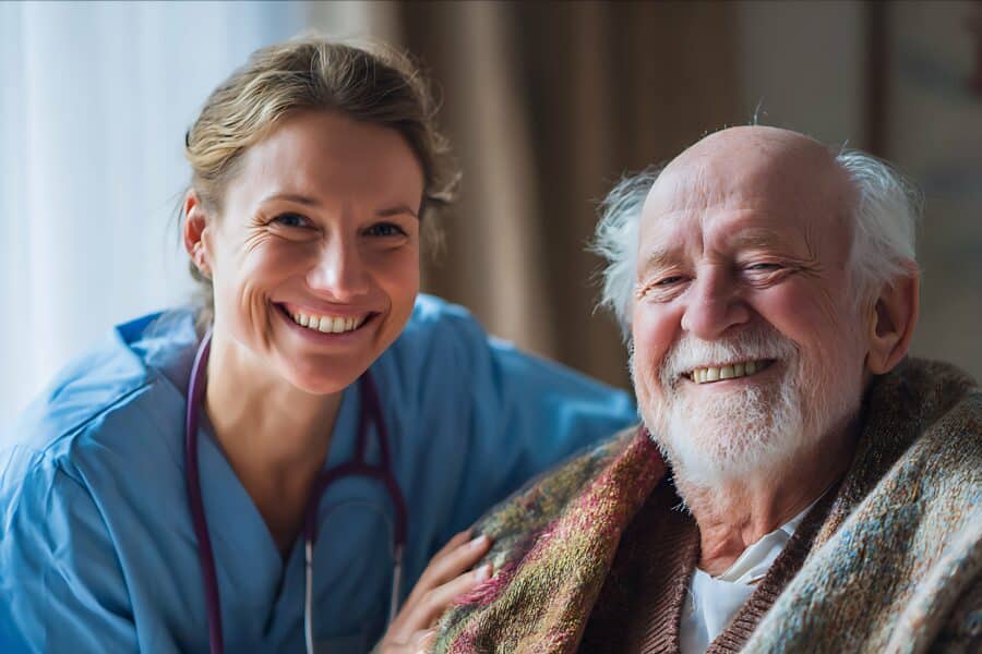 A smiling nurse in blue scrubs with stethoscope beside an elderly male care home resident during the Christmas period.
