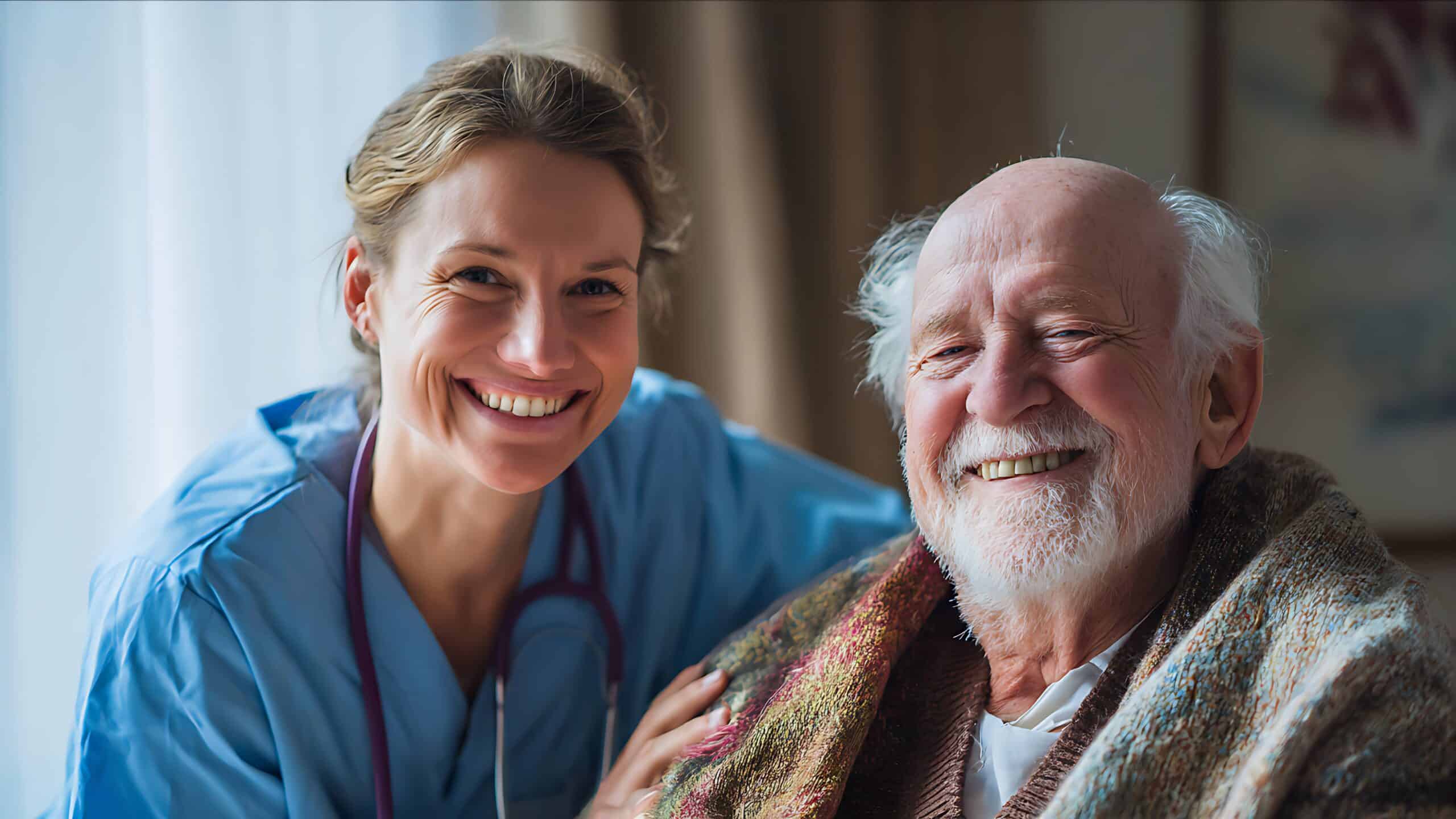 A nurse in scrubs smiles beside an elderly man wrapped in a blanket, both looking at the camera in a well-lit room.